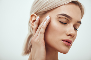 Beautiful young blonde woman keeping eyes closed and touching her face with pre-surgery markings